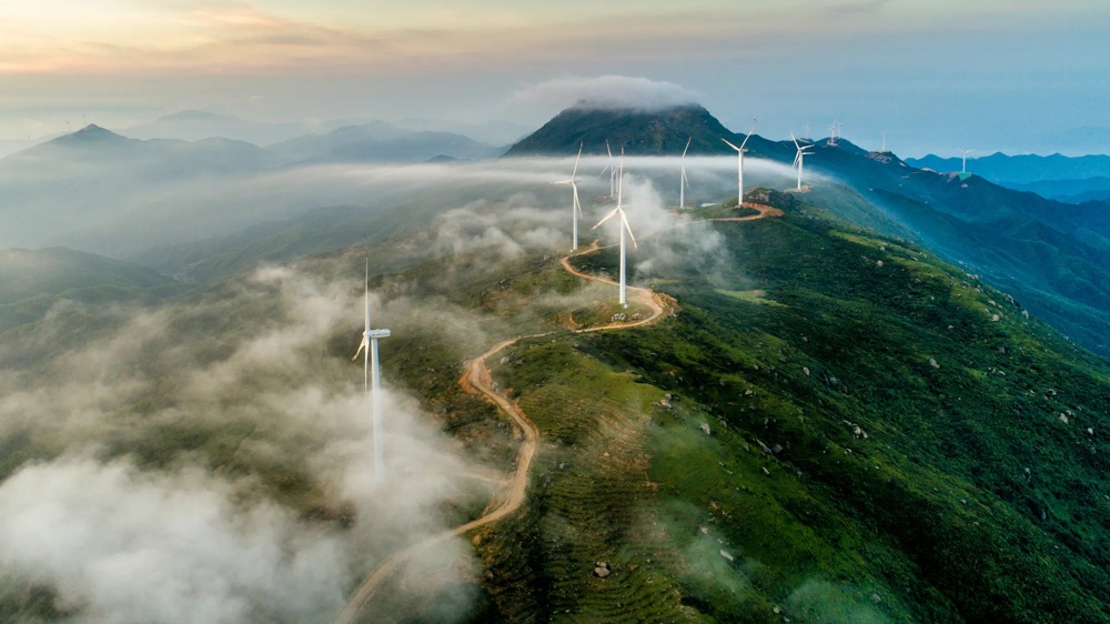 Windmills on a fogged mountain ridge at sunrise, surrounded by green landscape.