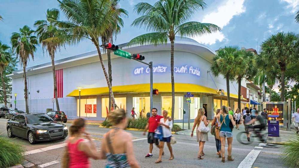 Menschen überqueren eine Straße vor einem Gebäude mit Palmen und gelbem Vordach in Miami Beach.