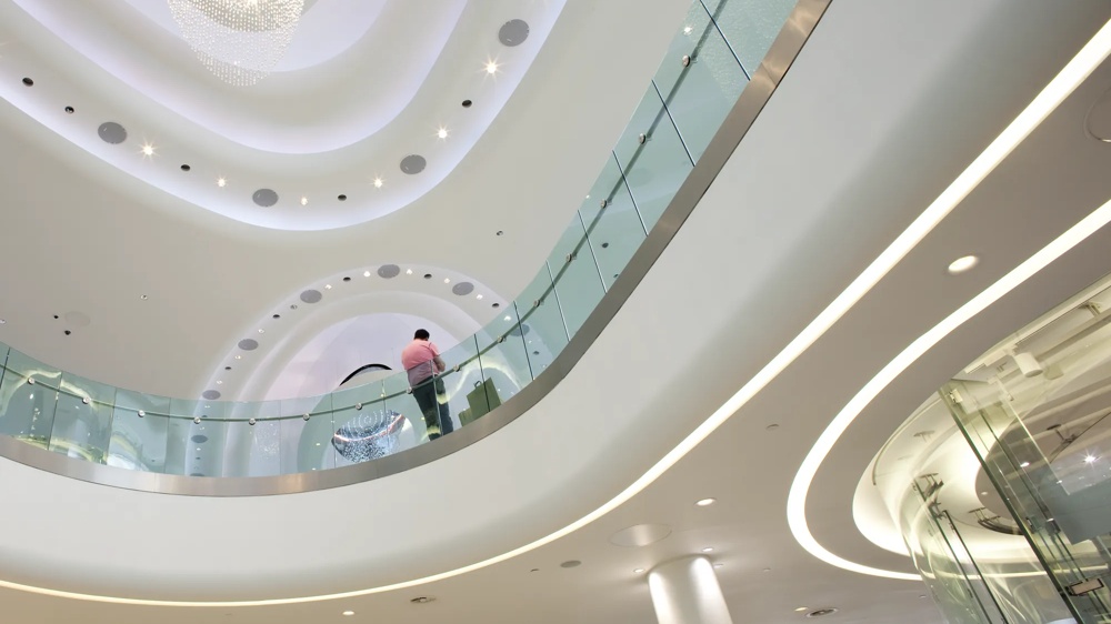 Moderne Innenarchitektur mit geschwungenen Decken, Glasgeländern und einem Kronleuchter. Eine Person in Pink steht oben.