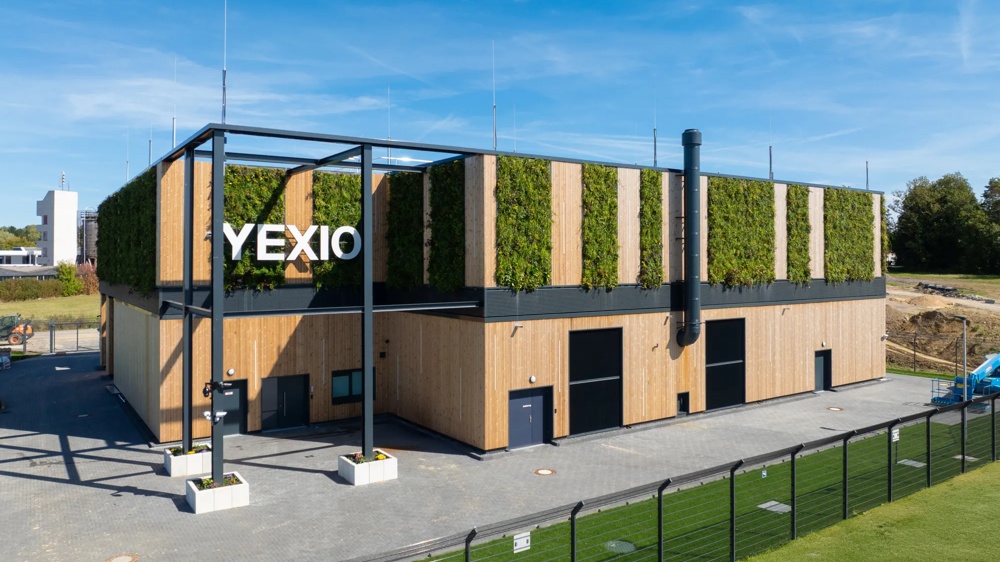 Ein modernes Gebäude mit Holzfassade und begrüntem Dach neben einem Zaun und einer Grünfläche.