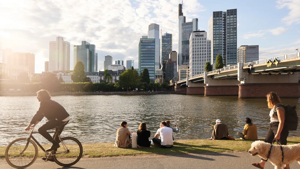 Personen sitzen am Flussufer, ein Radfahrer und eine Frau mit Hund im Vordergrund, Hochhäuser im Hintergrund.