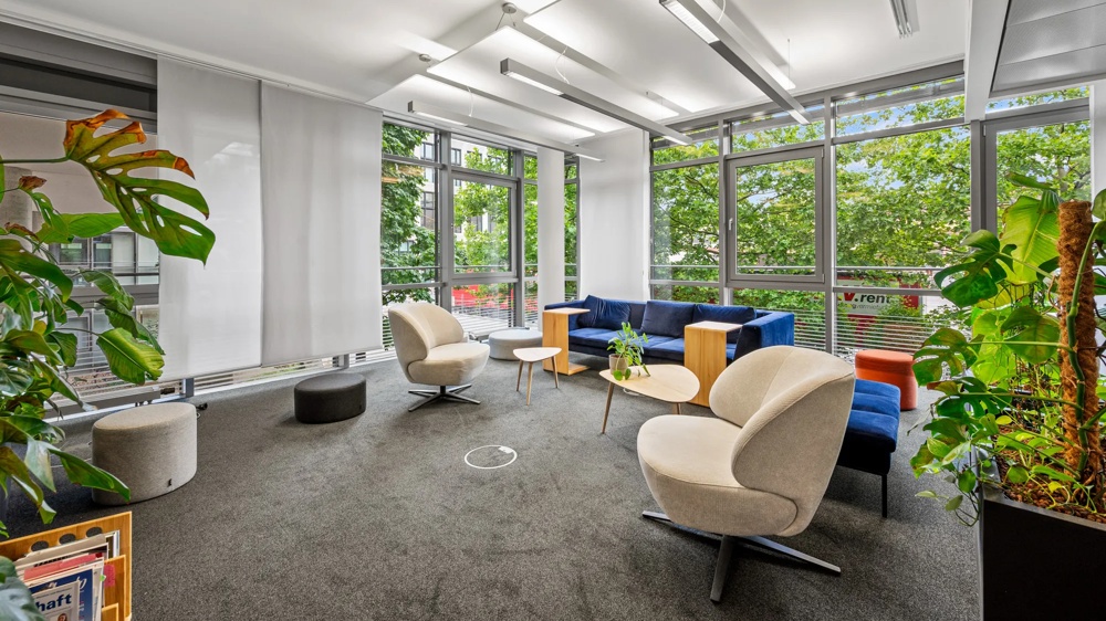 Ein moderner, heller Raum mit großen Fenstern, Pflanzen, blauen Sofas und beigen Sesseln auf grauem Teppich.