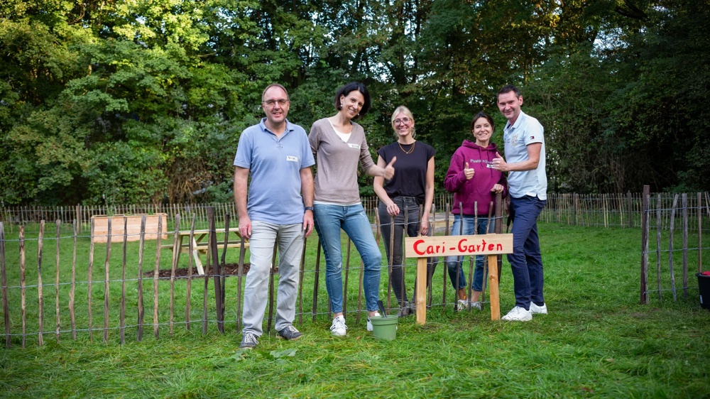 Five people are smiling behind a fence with a sign “Cari Garden” on a green meadow.