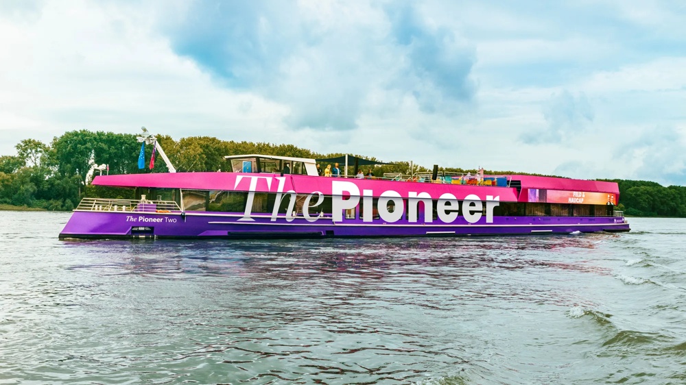 Ein pink-lila Boot mit der Aufschrift "The Pioneer" fährt auf einem Fluss, Bäume im Hintergrund.