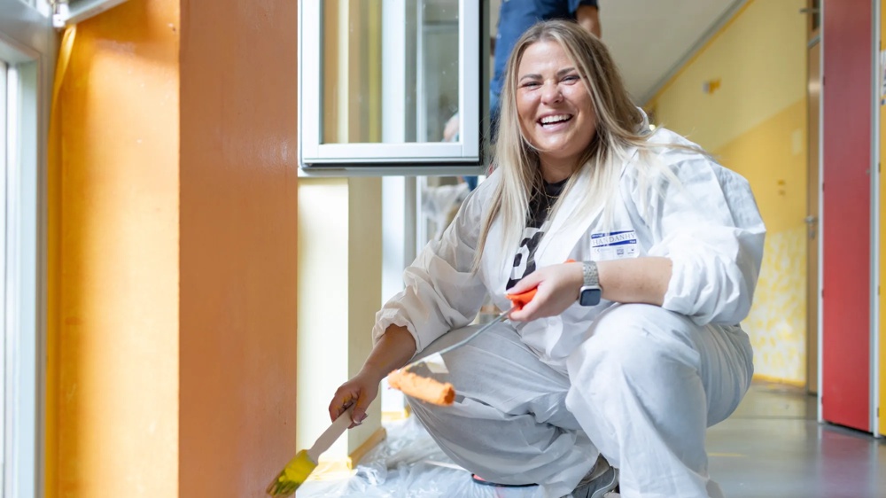 Frau in weißem Overall malt orange Wand, lächelt, kniet auf Plastikplane.