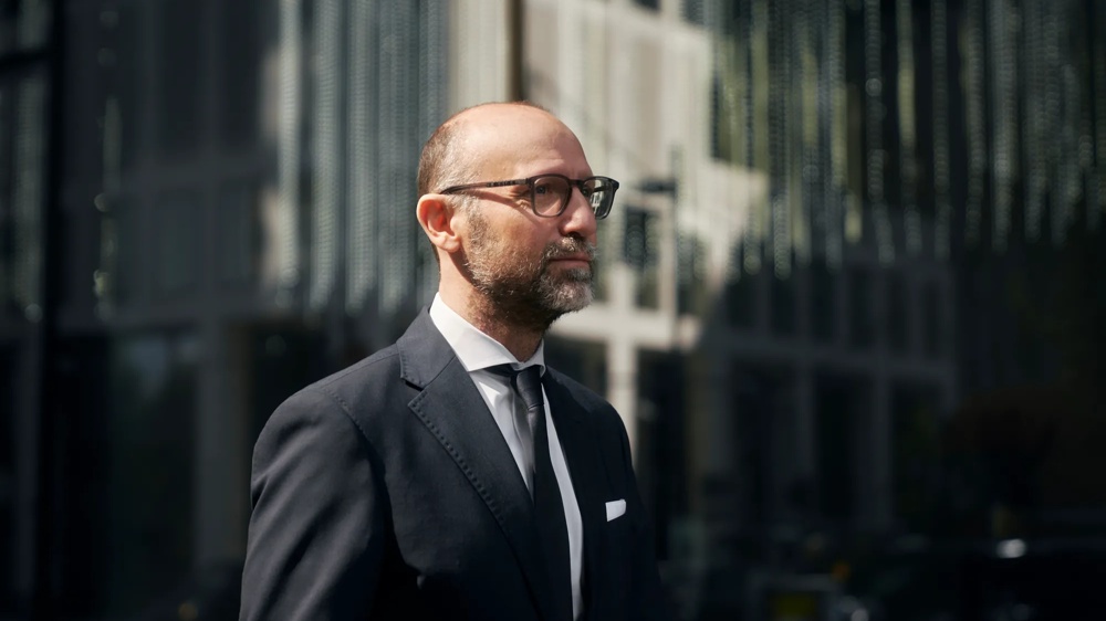 Ein Mann im Anzug mit Brille geht vor einem modernen Gebäude.