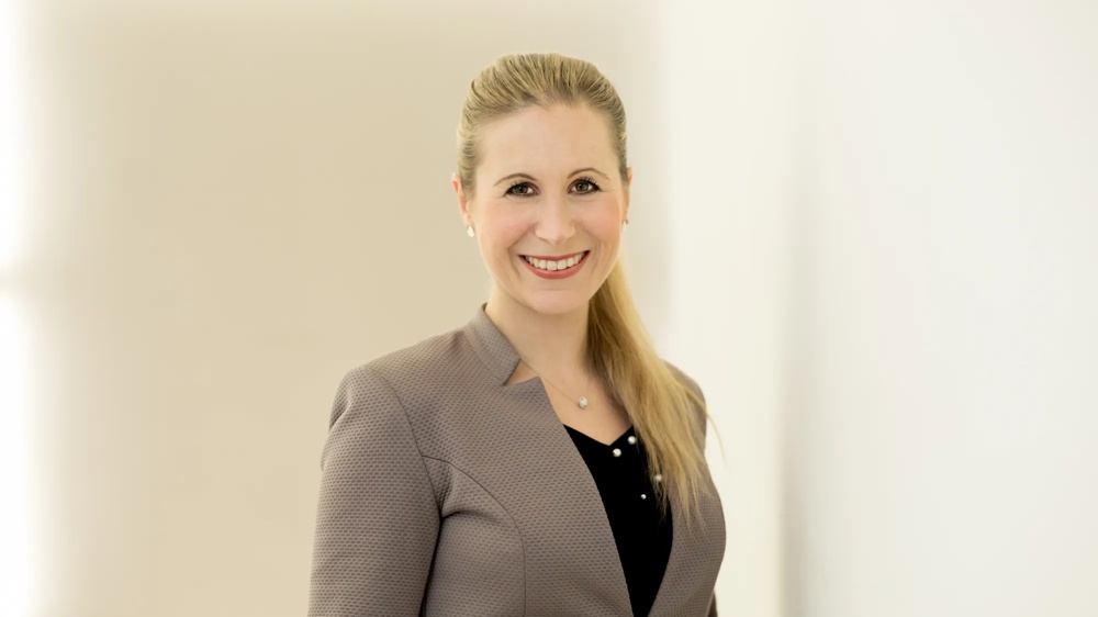 A smiling woman with a blonde ponytail in a grey blazer against a light background.