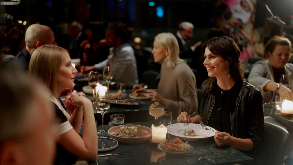 Zwei Frauen sitzen lächelnd bei Kerzenschein an einem Tisch in einem belebten Restaurant.