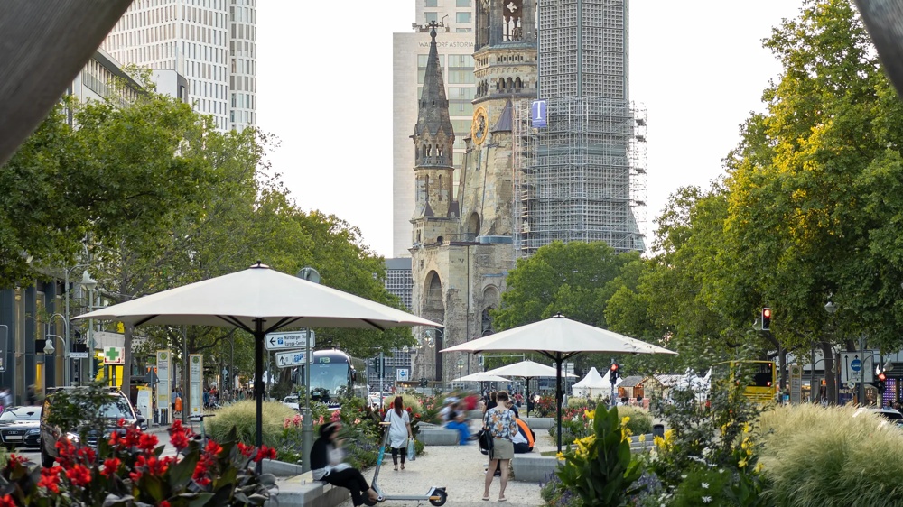 Ein Park mit Blumen, Sonnenschirmen und Menschen. Im Hintergrund eine teils eingerüstete Kirche.