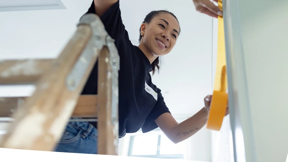 Person auf Leiter klebt gelbes Klebeband an Wand. Sie trägt ein schwarzes T-Shirt und lächelt.