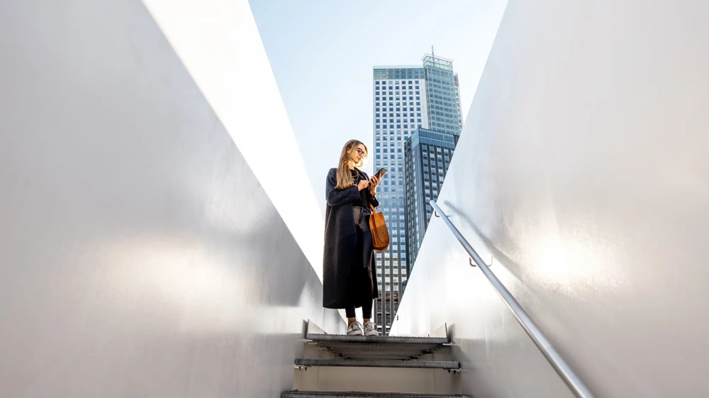 Woman with mobile phone on stairs, high-rise building in the background, blue sky.