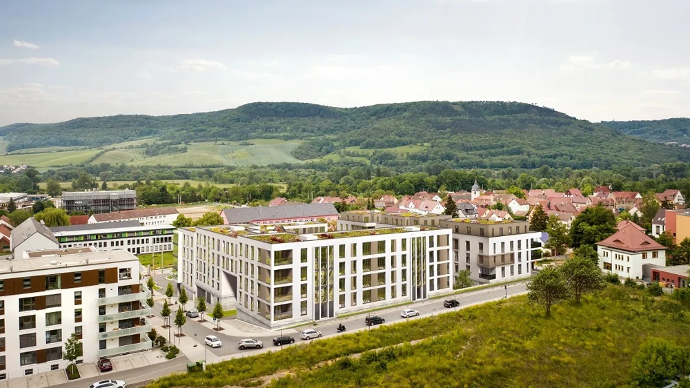 Moderne Gebäude vor grünen Hügeln und einem Dorf mit roten Dächern unter bewölktem Himmel.
