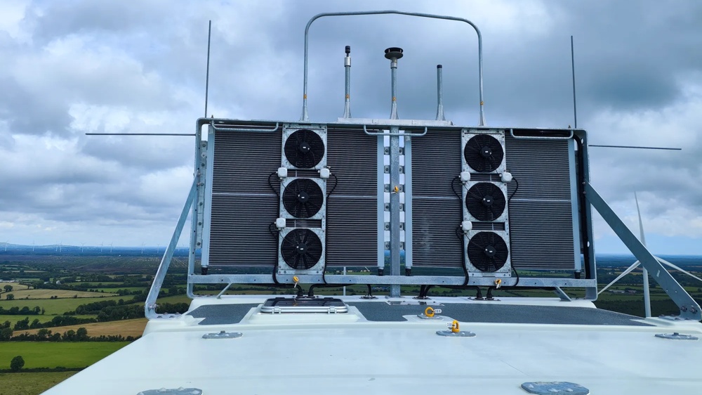 Technische Kühleinheit mit mehreren Lüftern auf dem Dach einer Windenergieanlage vor einer weiten Landschaft und einem bewölkten Himmel.