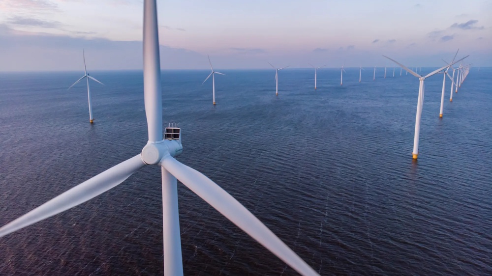 Windpark im Meer bei Sonnenuntergang, mit mehreren Windturbinen in einer Reihe über ruhigem Wasser.