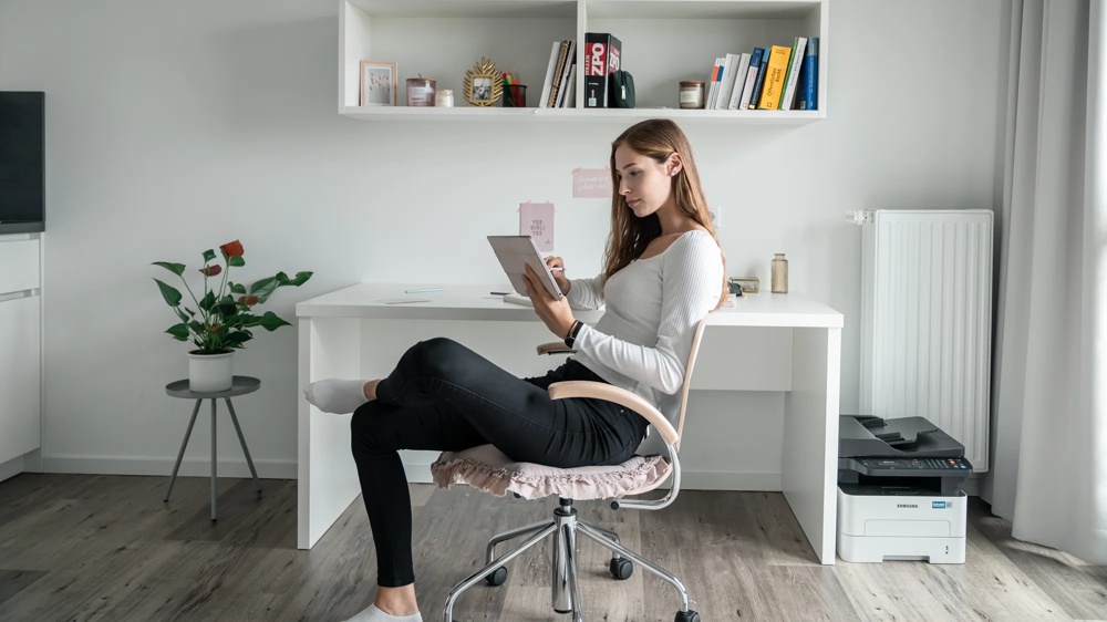 Frau sitzt an weißem Schreibtisch, liest Tablet; Bücherregal, Pflanze und Drucker im Hintergrund.