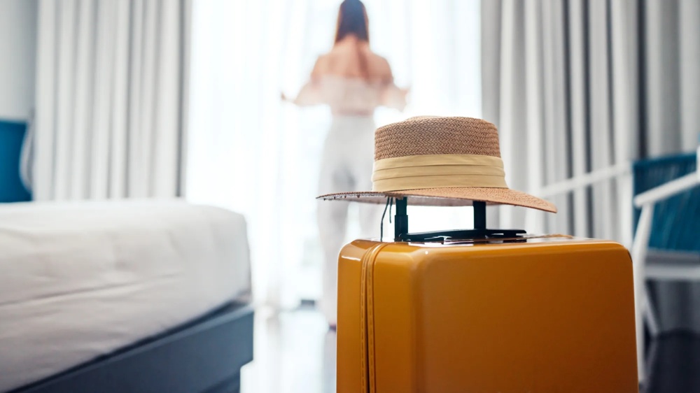 A straw hat is lying on a yellow suitcase in a bright room with a person by the window.