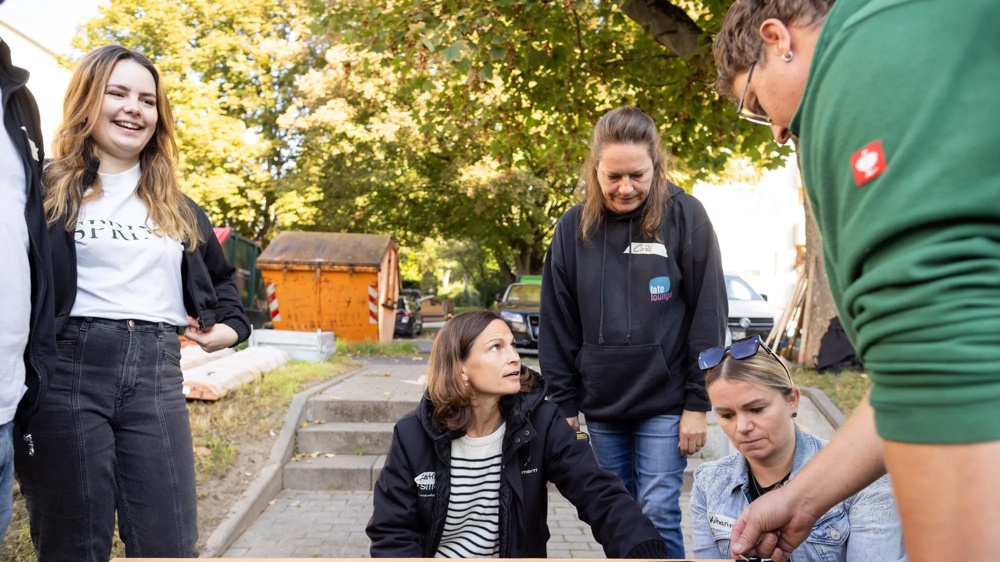 A group of five people standing and kneeling around a black wooden frame outdoors, surrounded by trees.
