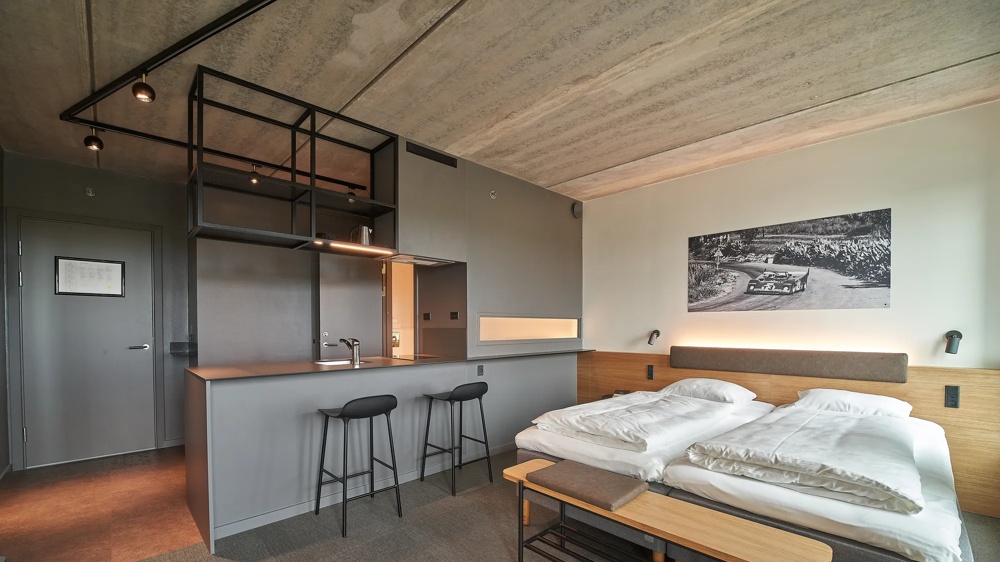 Modern hotel room with double bed, grey kitchenette, two bar stools and black and white photo on the wall.