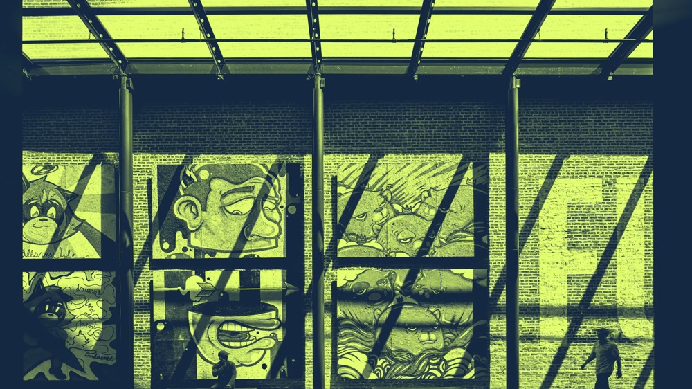 Wandgemälde mit Cartoon-Gesichtern in einer Halle, zwei Personen davor, Schatten von Glasdach fallen auf die Wand.