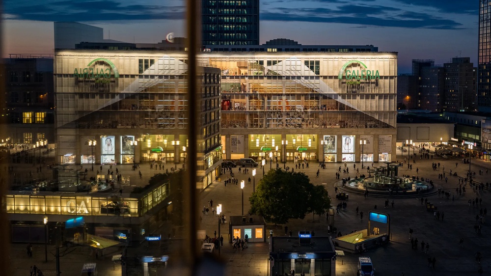 Am Berliner Alexanderplatz wird die Fassade der Galeria-Filiale gezeigt, wie sie von Fassadenkletterern mit bedruckten Klebebahnen versehen wird. die Bahnen visualisieren die mögliche neue Innengestaltung des Gebäudes als offene Bibliothek.