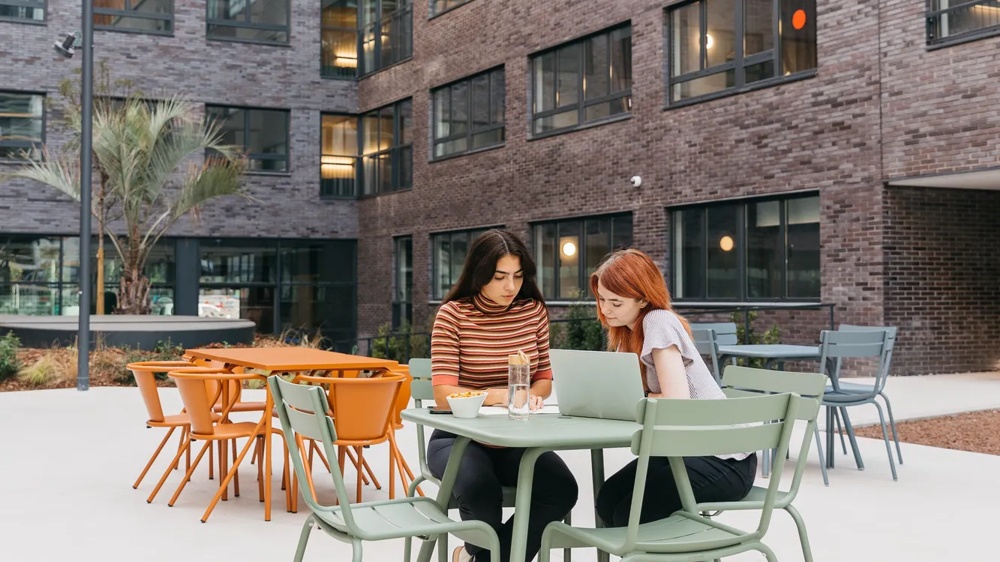Zwei Frauen sitzen an einem grünen Tisch im Innenhof eines modernen Gebäudes und arbeiten an einem Laptop.