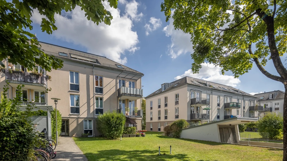 Mehrfamilienhäuser mit Balkonen, grüner Rasen, Bäume und Fahrräder im Vordergrund unter blauem Himmel.