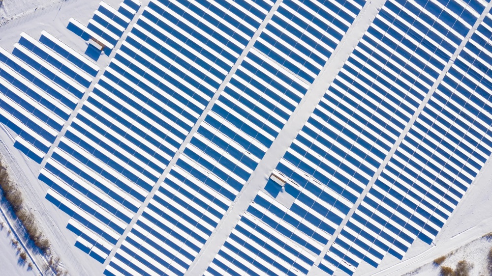 Luftaufnahme einer verschneiten Solaranlage mit blauen Paneelen in gleichmäßigen Reihen.