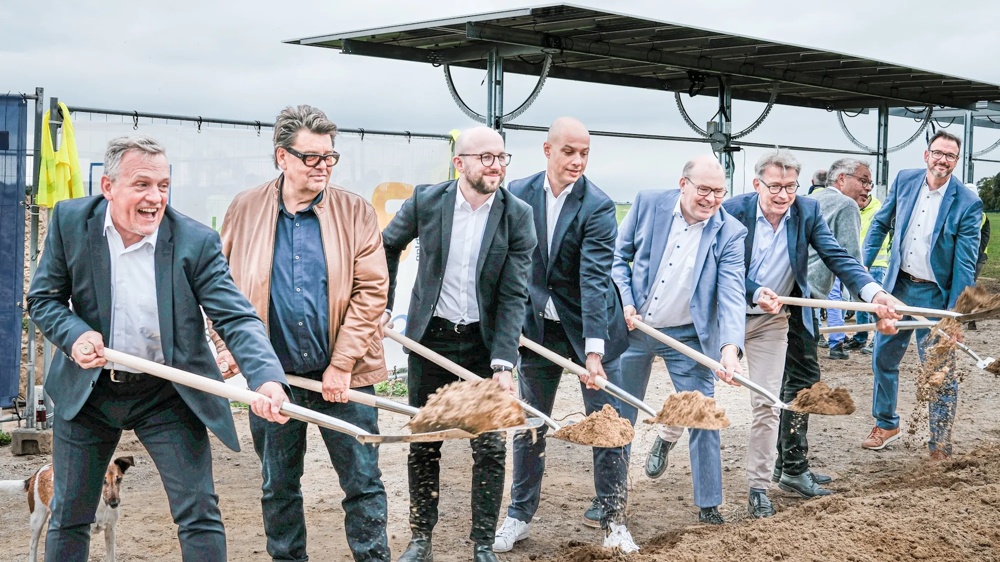 Acht Männer in Anzügen heben mit Schaufeln Erde auf einer Baustelle.