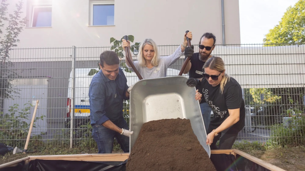 Five people tipping soil from a wheelbarrow into a raised bed outdoors.