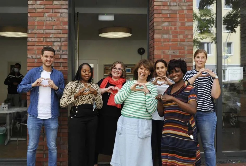 A group of eight people standing in front of a building smiling and shaping heart symbols with their hands.