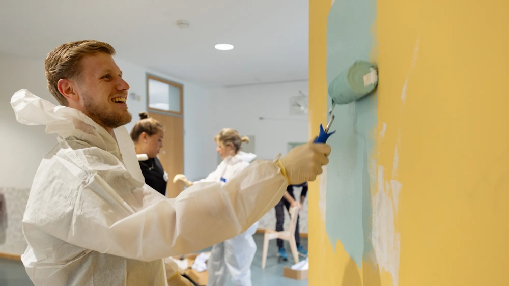 Person in protective suit painting a yellow wall with blue paint, other people in the background.