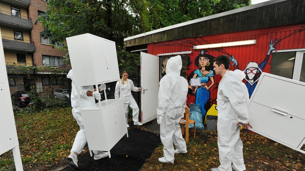Personen in weißen Schutzanzügen tragen weiße Schränke vor ein rot bemaltes Gebäude.
