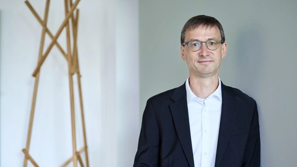 Ein Mann mit Brille im Anzug steht vor einer grauen Wand und schaut freundlich in die Kamera.