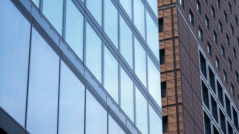 Zwei moderne Hochhäuser, eines mit Glasfassade, das andere mit brauner Ziegeloptik, stehen nebeneinander.