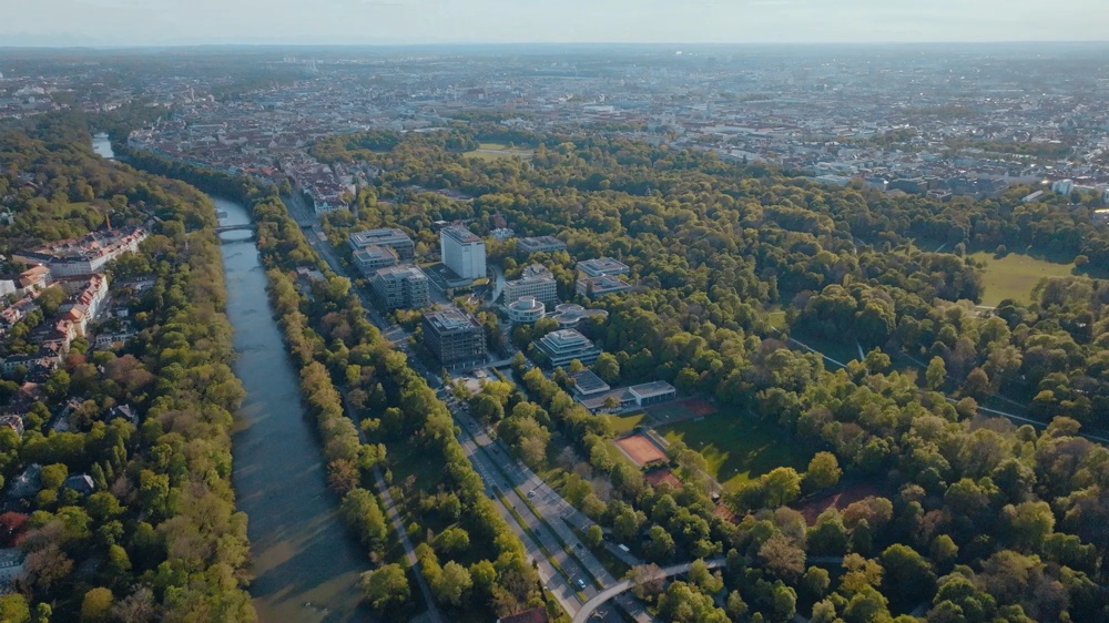 Aerial shot of a city with a river, surrounded by dense, green trees and buildings in the background.