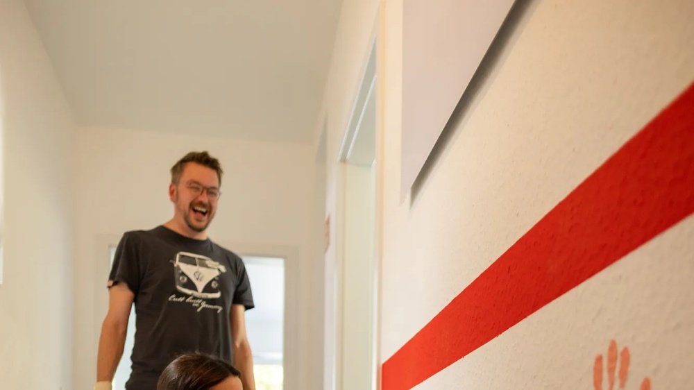 Woman pressing handprint in orange on white wall, surrounded by paint cans in the hallway.