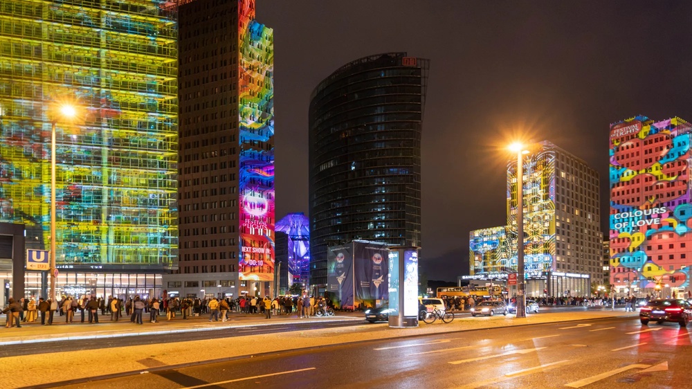 Mehrgeschossiges Gebäudeensemble bei Nacht mit großflächigen farbigen Lichtprojektionen auf den Fassaden.
