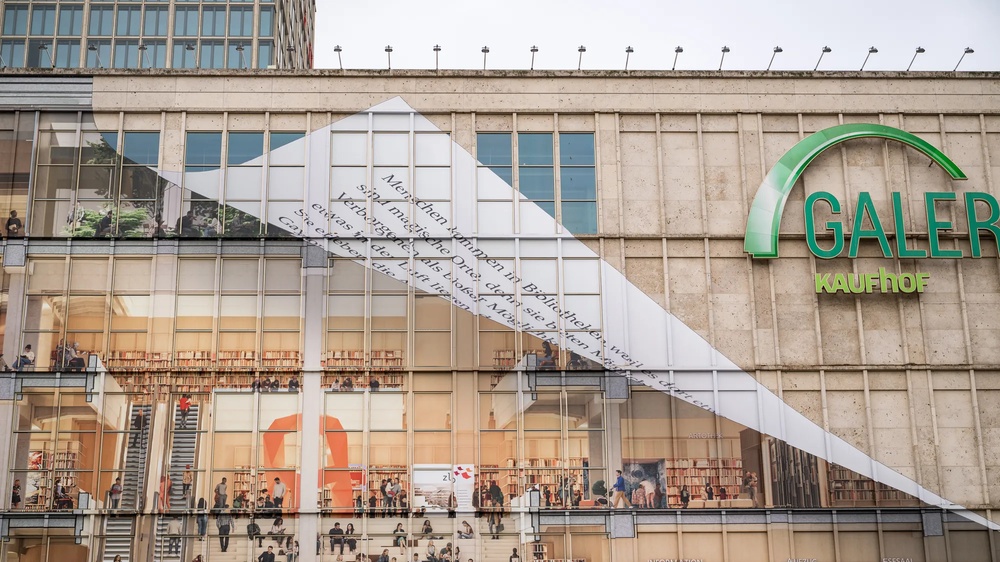 Am Berliner Alexanderplatz wird die Fassade der Galeria-Filiale gezeigt, wie sie von Fassadenkletterern mit bedruckten Klebebahnen versehen wird. die Bahnen visualisieren die mögliche neue Innengestaltung des Gebäudes als offene Bibliothek.