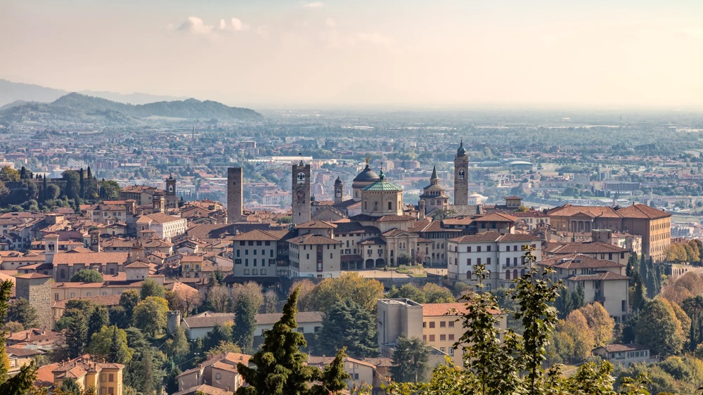 Panorama der Altstadt von Bergamo mit roten Ziegeldächern, historischen Türmen und sanften Hügeln im Hintergrund.