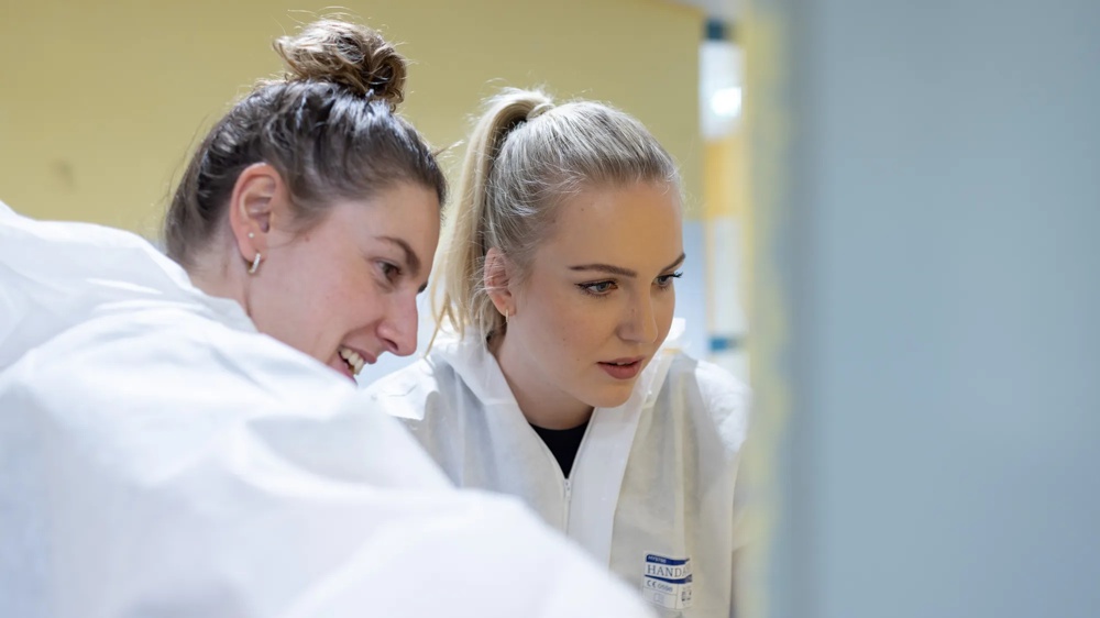 Zwei Frauen in weißen Schutzanzügen schauen konzentriert auf einen Bildschirm.