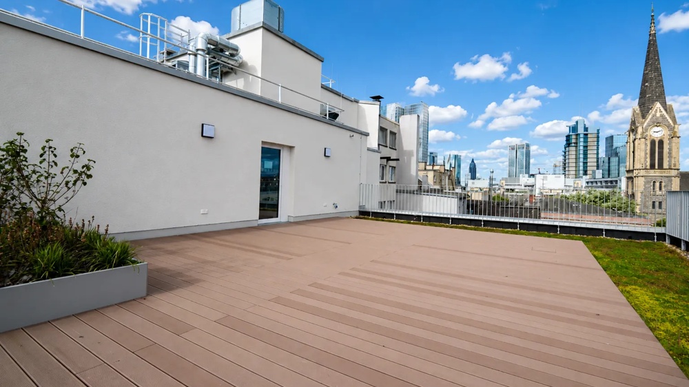 Dachterrasse der Büroimmobilie Schäfergasse 33 in Frankfurt am Main mit Holzdeck und Begrünung, im Hintergrund die Frankfurter Skyline mit Kirche und Bankentürmen – ein hausInvest-Objekt.