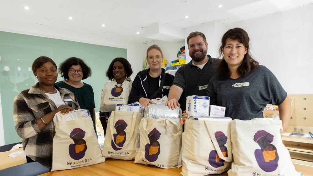 Six people are smiling behind fabric bags with care products at a wooden table.