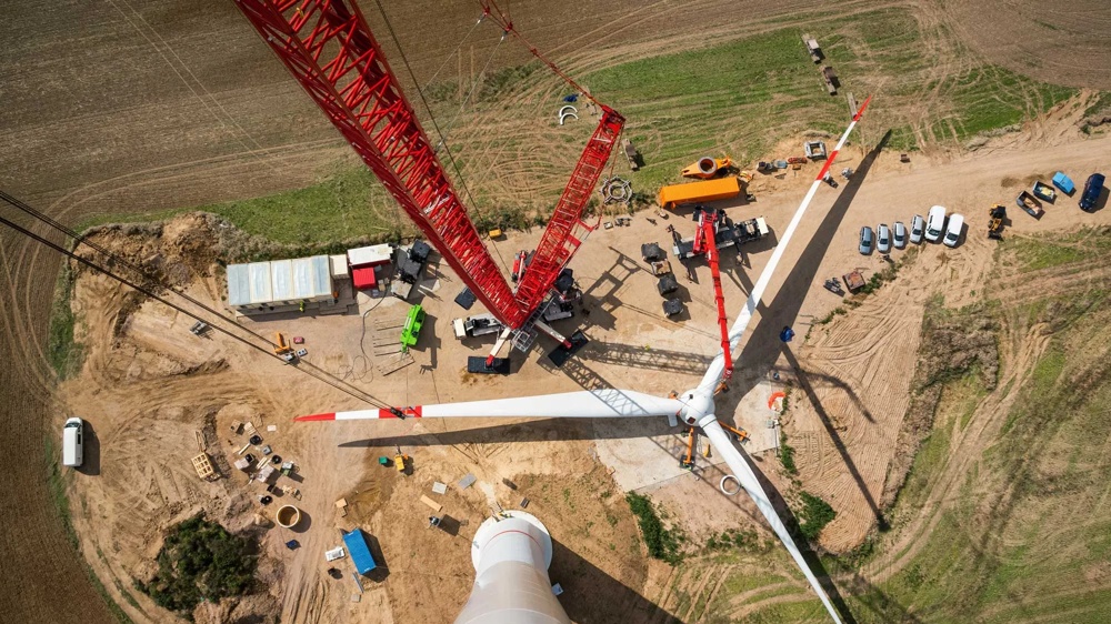 Blick von oben auf eine Windkraftanlage im Bau, mit rotem Kran und Fahrzeugen auf einem Feld.