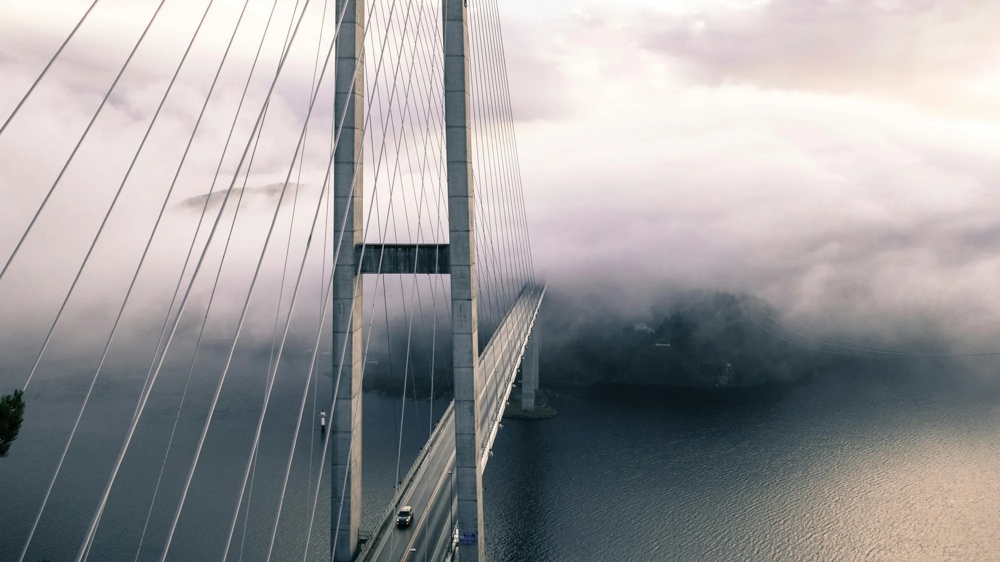 Eine lange Hängebrücke führt über nebliges Wasser, umgeben von dichtem Nebel.