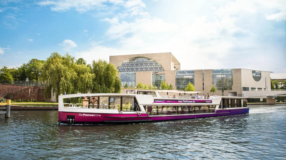 Ein modernes Ausflugsboot fährt auf einem Fluss vor einem großen, modernen Gebäude und grünen Bäumen.