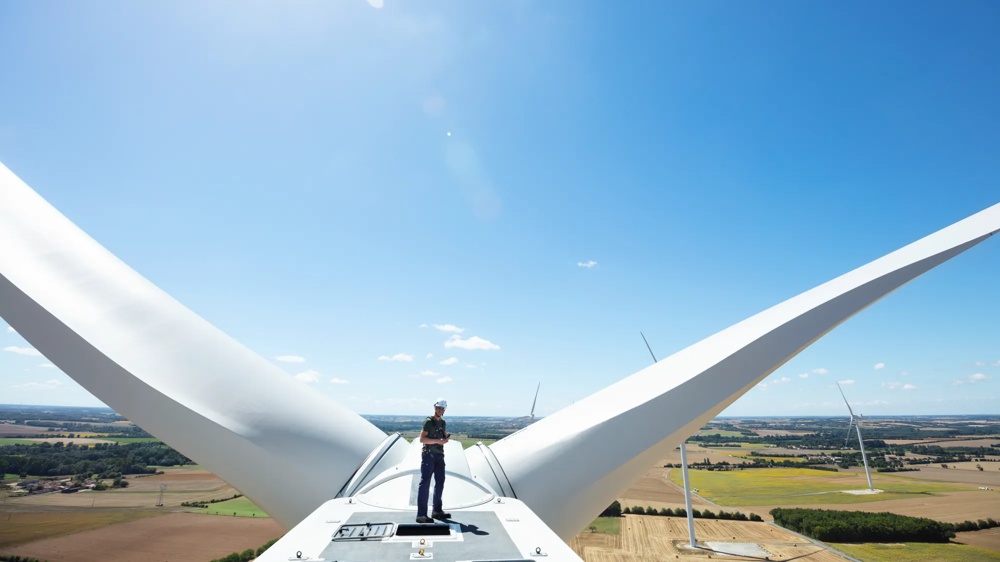 Person steht auf Windradnabe, blauer Himmel, weite Landschaft im Hintergrund.