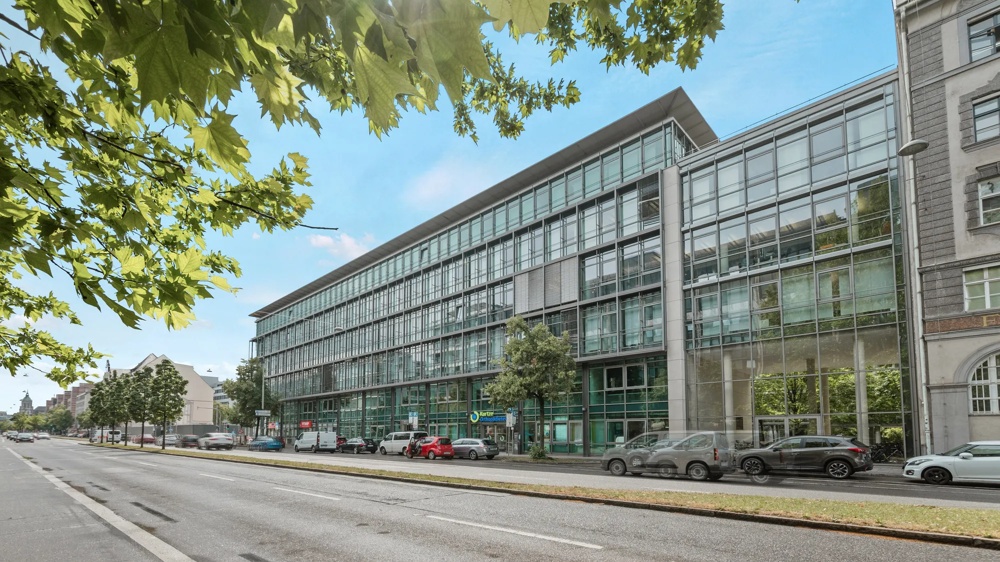 Moderne Glasfassade eines Bürogebäudes an einer belebten Straße, Bäume im Vordergrund, blauer Himmel.