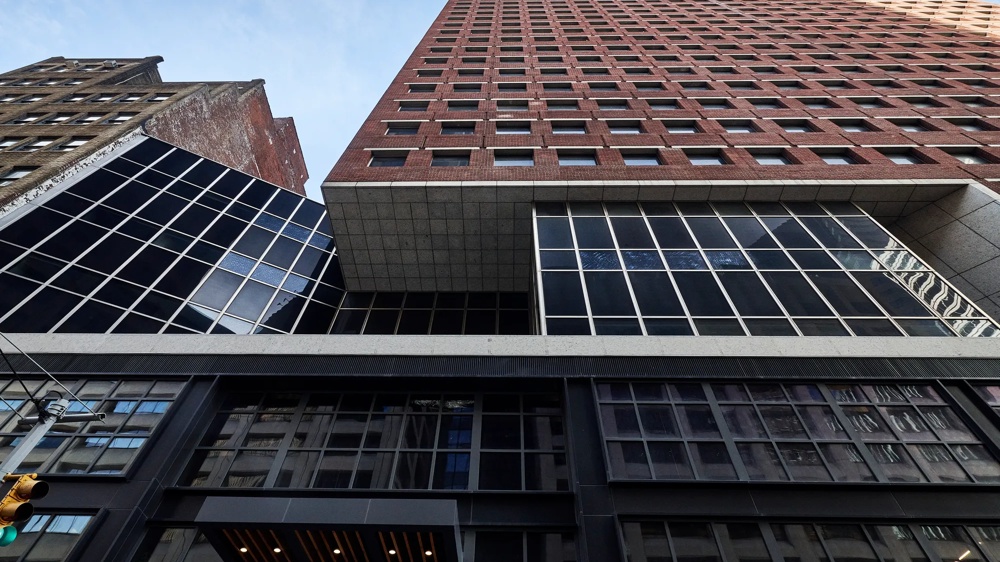 Blick nach oben auf ein hohes Gebäude mit roter Ziegelfassade und großen Glasfenstern, darunter ein älteres Gebäude.
