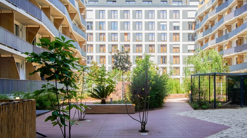 Courtyard with modern residential buildings, balconies, green plants and a central walkway under a blue sky.