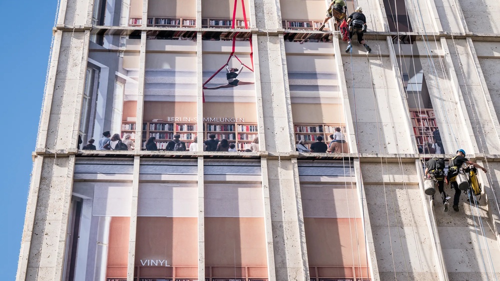 Am Berliner Alexanderplatz wird die Fassade der Galeria-Filiale gezeigt, wie sie von Fassadenkletterern mit bedruckten Klebebahnen versehen wird. die Bahnen visualisieren die mögliche neue Innengestaltung des Gebäudes als offene Bibliothek.